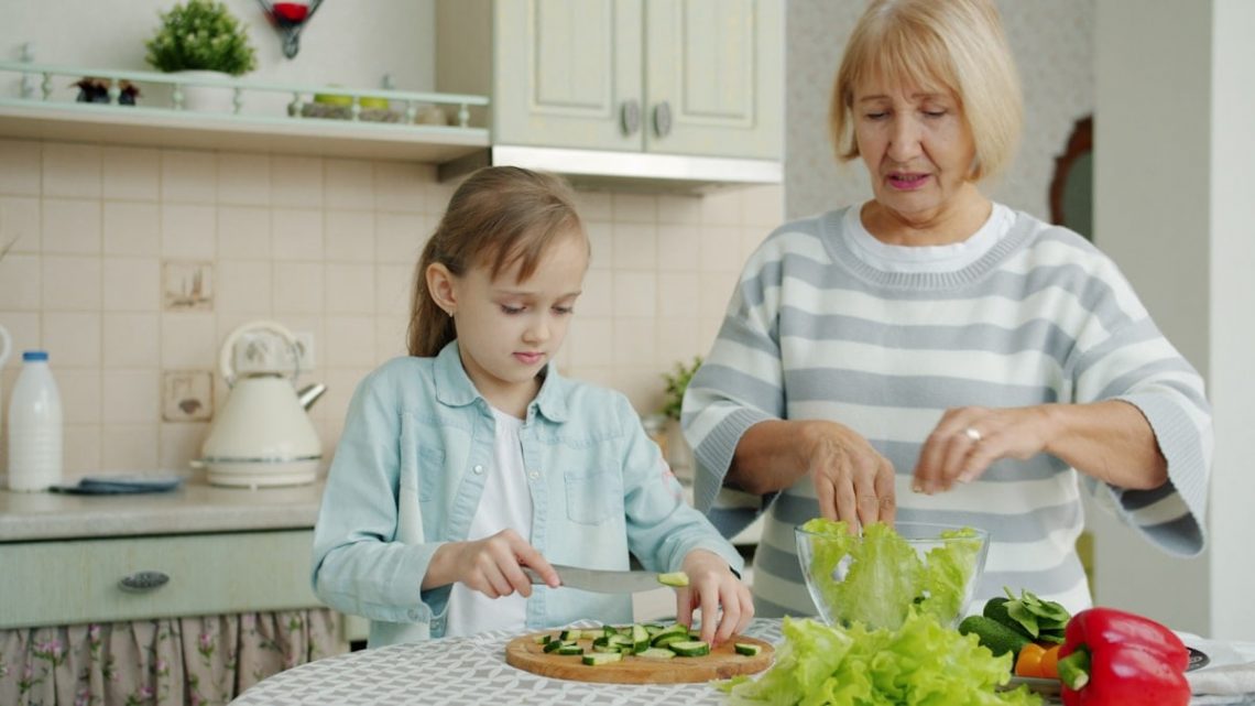 Grandma and cute child are cooking salad for lunch talking making healthy meal at home. Domestic activities, family life and happy childhood concept.
