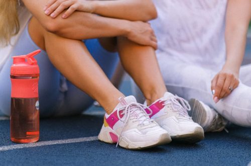 Crop anonymous female friends in activewear sitting on sports ground after training near bottle with water in daytime