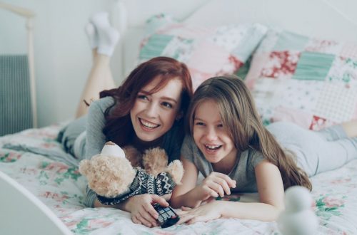 Laughing happy mother and cute daughter watching funny cartoon movie on TV while lying on bed at home in morning have fun at cozy bedroom